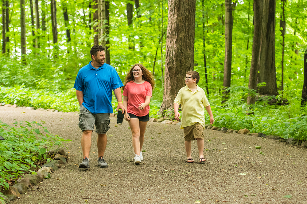 Older man in blue polo walking alongside young girl in pink shirt and young boy in yellow shirt on a trail in a wooded area