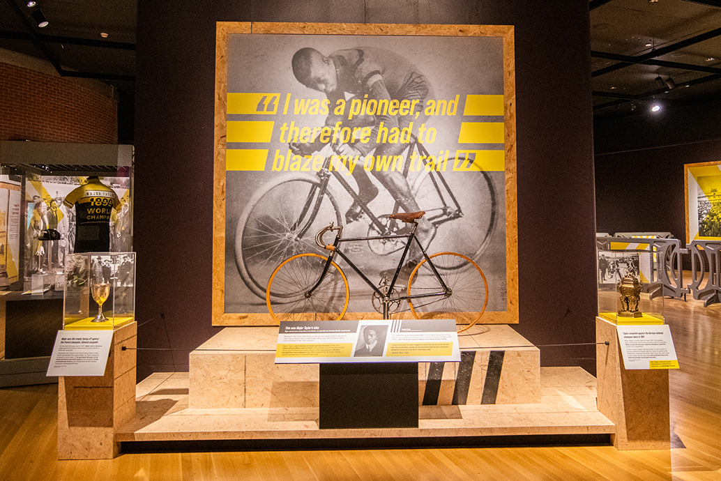 Black and white photo of Major Taylor sitting on bike with yellow text over the top of it