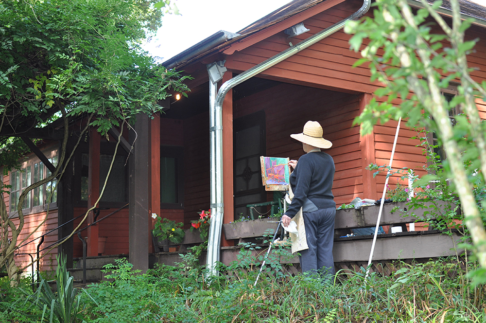 Woman wearing straw hat painting en plein air in front of red house