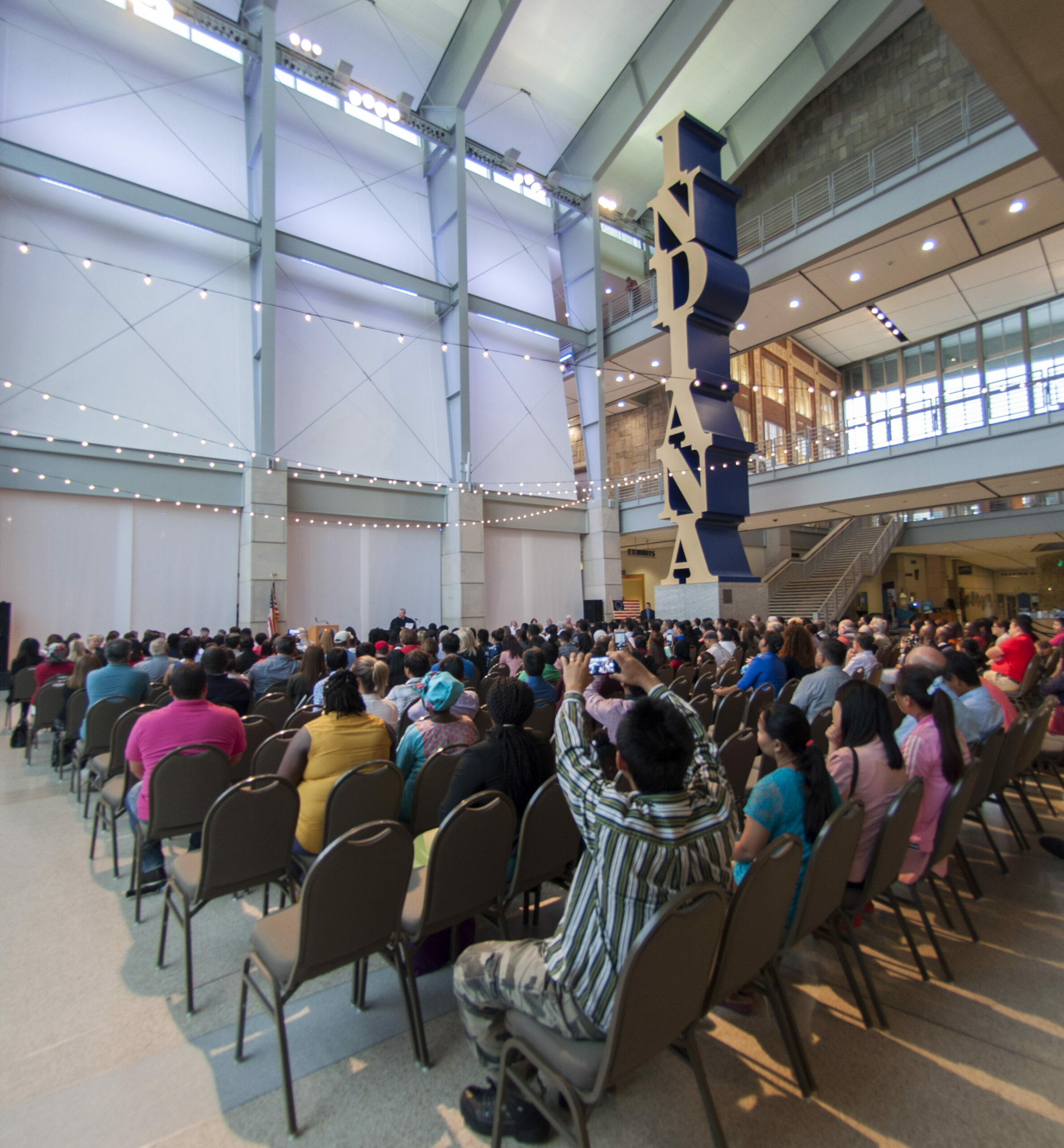 hundreds of people sitting in chairs in the Great Hall in front of the Indiana obelisk at the Indiana State Museum