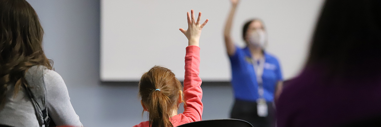 Girl in pink shirt raising hand in classroom in front of teacher