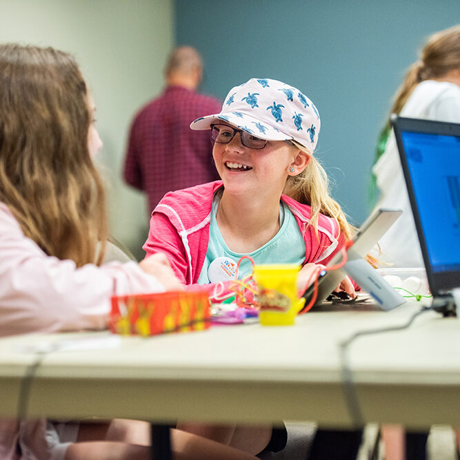 young girl with hat on, sitting at table smiling